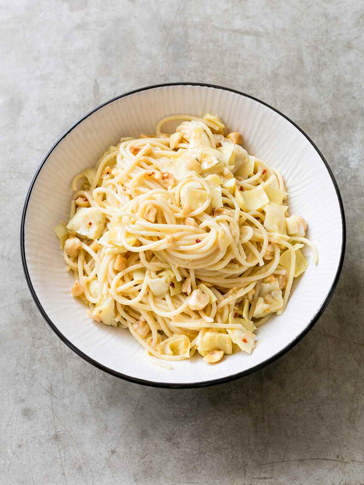 En stor vit skål fylld med spagetti, bitar av kronärtskockor, hasselnötter, fläckar av chilipepparflingor och parmesanost.