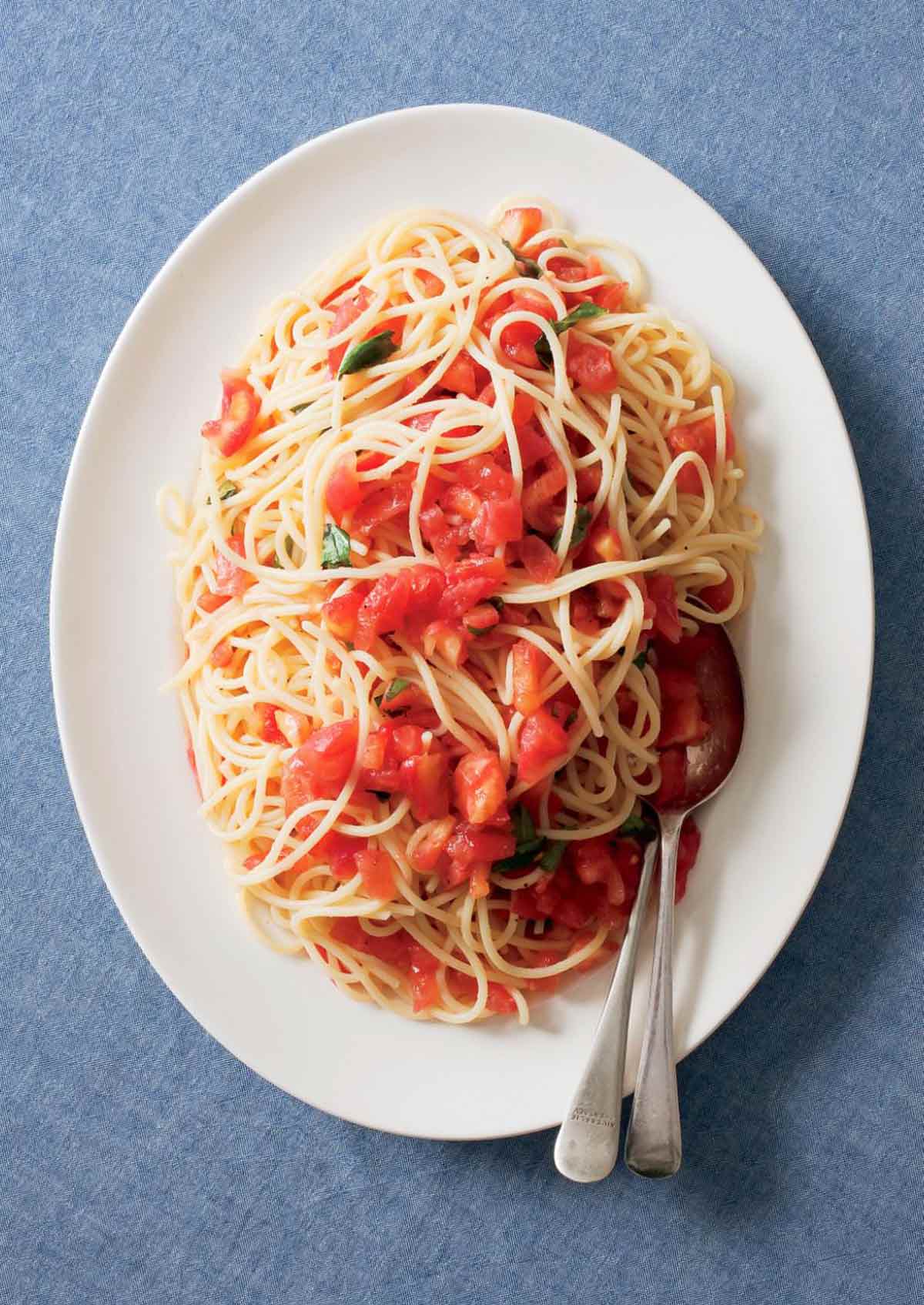 Ett ovalt fat toppat med spagetti med råa tomater och ett par skedar vid sidan av.