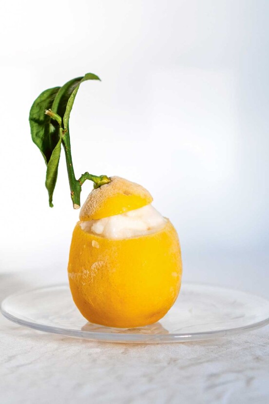 A frosty, hollowed out lemon wih leaf, filled with pale lemon sorbet on a glass plate.
