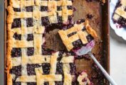 A large cookie sheet filled with blueberry slab pie with a lattice top, with pieces missing and a spatula.