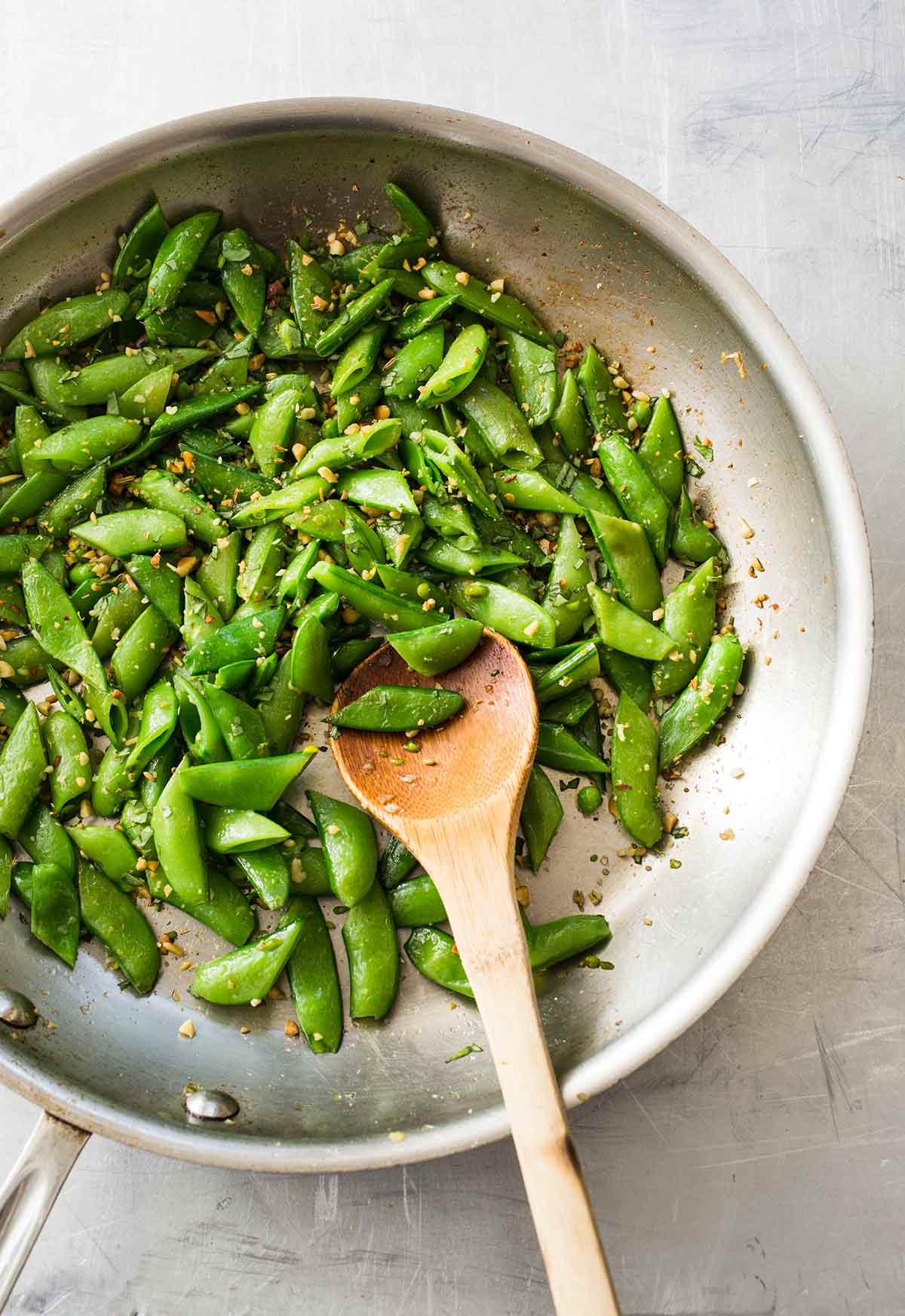 A metal wok filled with halved snap peas, sprinkled with fennel, pine nuts, and lemon zest, with a wooden spoon.