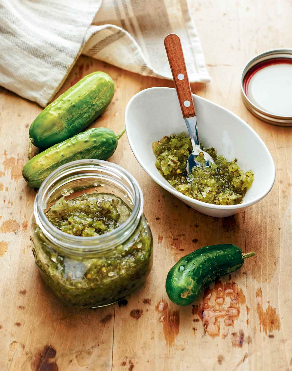 A jar and bowl of sweet pickle relish with three cucumbers lying beside them.