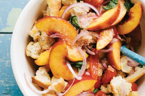 A blue background with a white bowl filled with sliced peaches, tomatoes, red onions, basil, and chunks of bread.