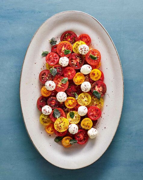 A large white platter filled with halved various colored cherry tomatoes, labneh balls, olive oil, and a sprinkle of spices.