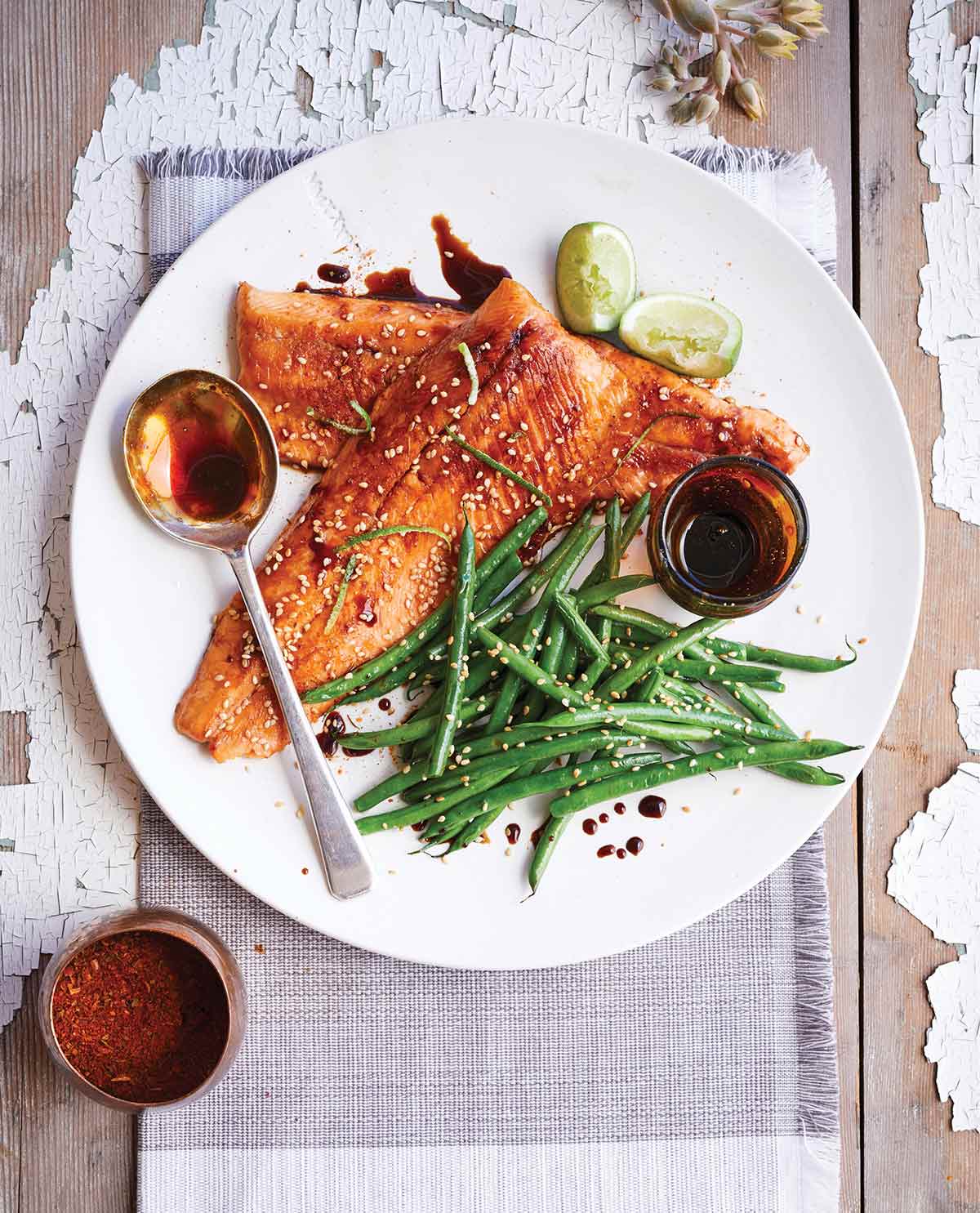 A weathered wood table with a white plate, filled with 2 honey-soy covered trout, green beans, lime quarters, a pot of glaze and a large spoon.