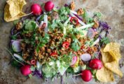 A pile of turkey taco salad piled up on a stainless steel sheet, with radishes, lettuce, cabbage, cilantro, and tortilla chips.