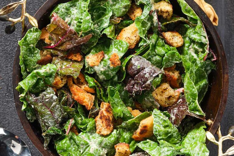 A dark bowl with handles full of Swiss chard Caeser salad and croutons on a dark background.