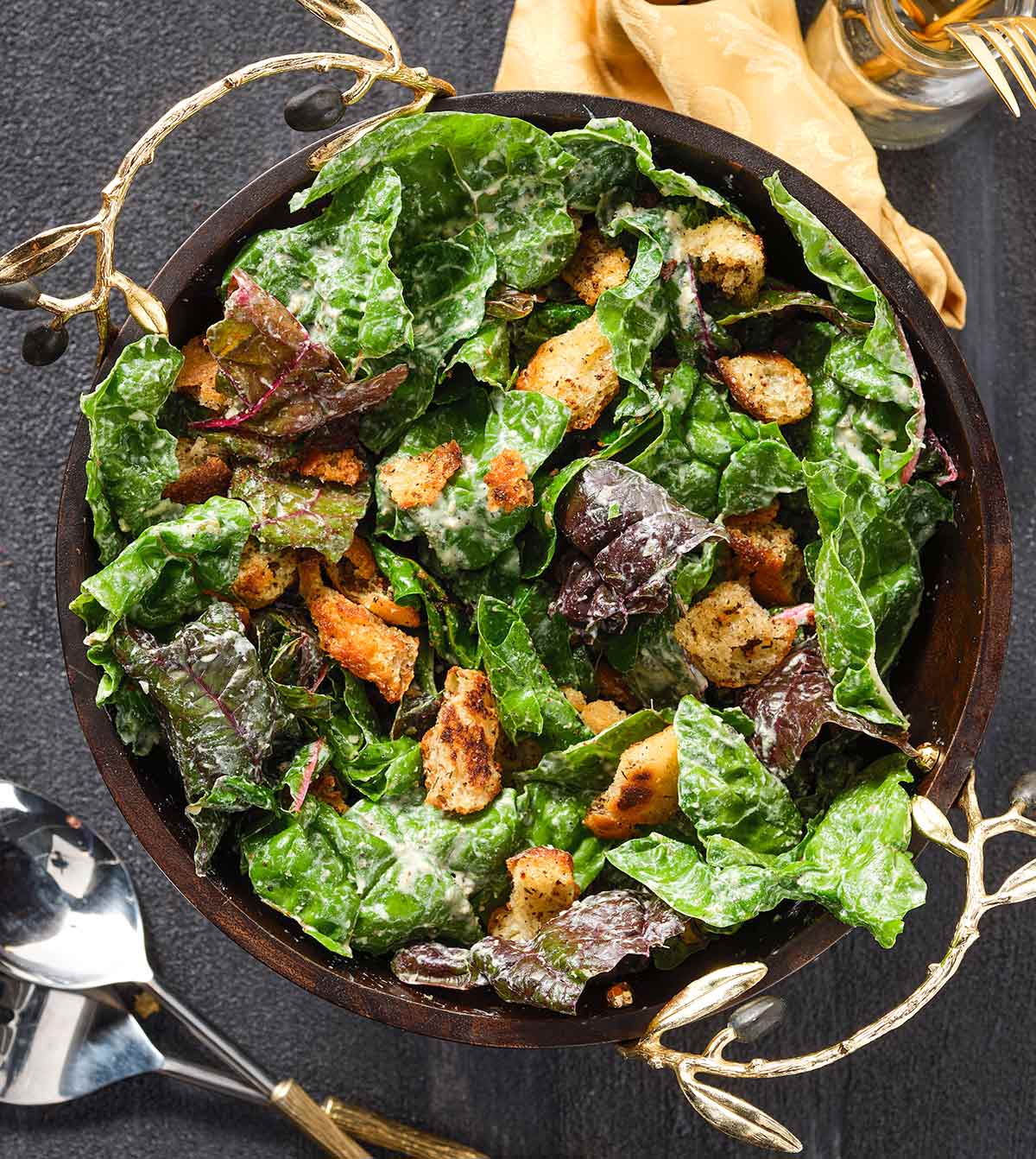 A dark bowl with handles full of Swiss chard Caeser salad and croutons on a dark background. 