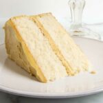 A slice of 2 layer white chocolate whisper cake with pistachio frosting and filling, on a white plate beside a cake stand containing the rest of the cake.