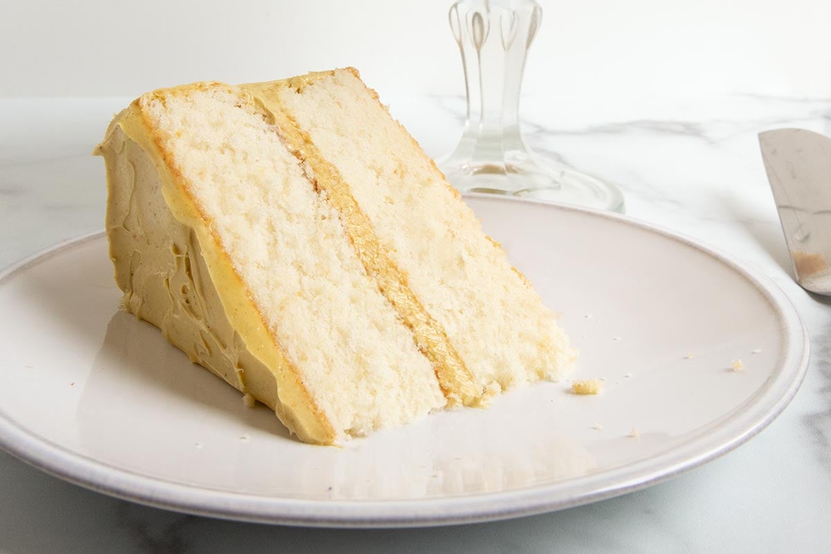 A slice of 2 layer white chocolate whisper cake with pistachio frosting and filling, on a white plate beside a cake stand containing the rest of the cake.