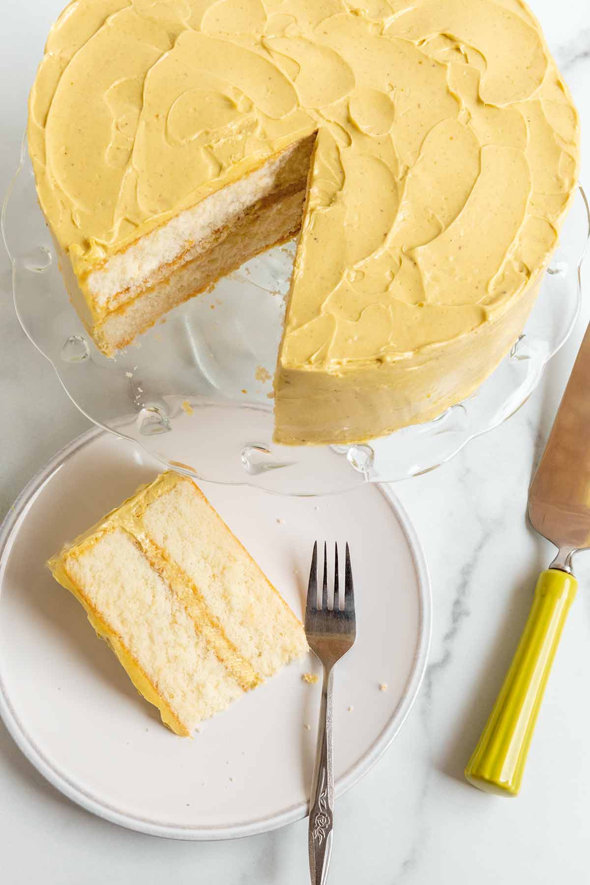 A slice of 2 layer white chocolate whisper cake with pistachio frosting and filling, on a white plate beside a cake stand containing the rest of the cake.