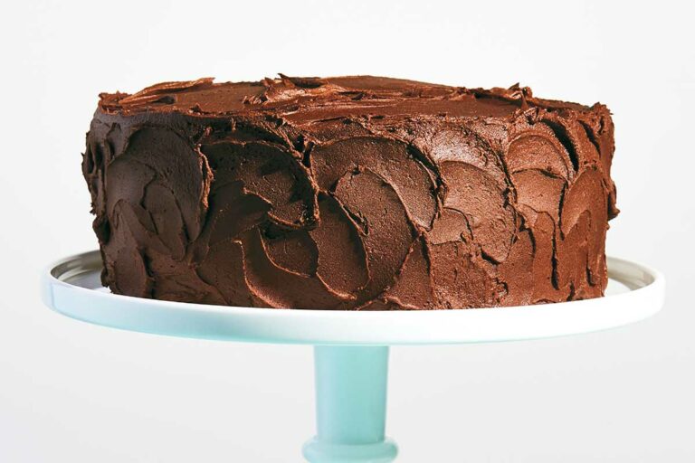 A yellow cake with chocolate frosting on a cake stand with a stack of plates beside it.