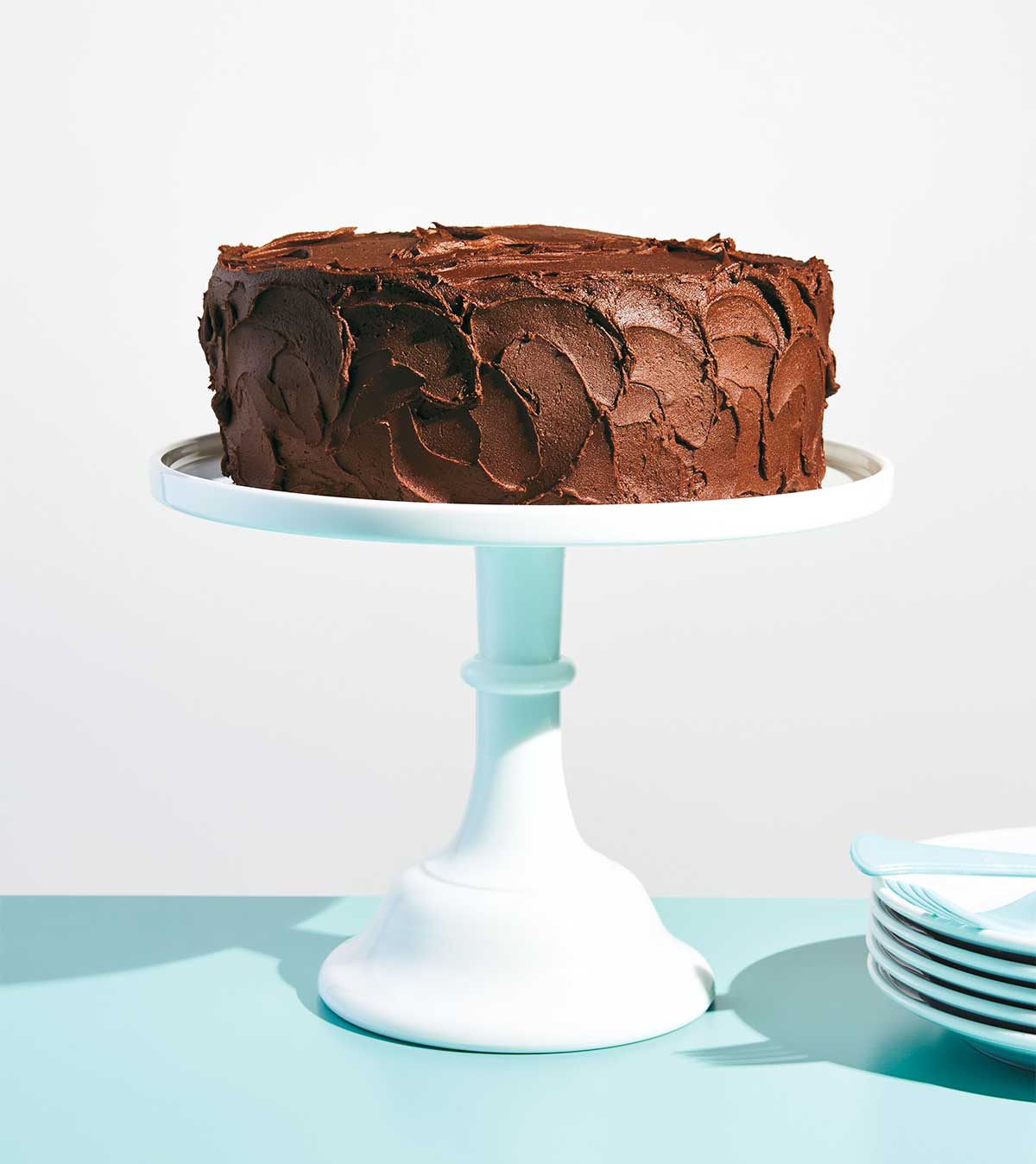 A yellow cake with chocolate frosting on a cake stand with a stack of plates beside it.