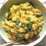 A green bowl filled with scrambled eggs and green pieces of zucchini on a tile tabletop, with a fork.