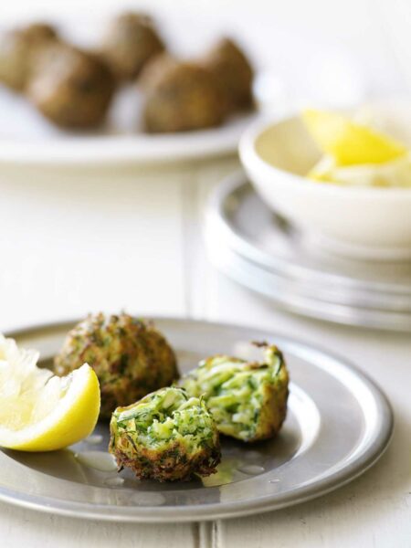A metal plate with 2 zucchini fritters on it, one broken in half. A slice of lemon lays beside them, a bowl of lemon and a plate of fritters in the background.