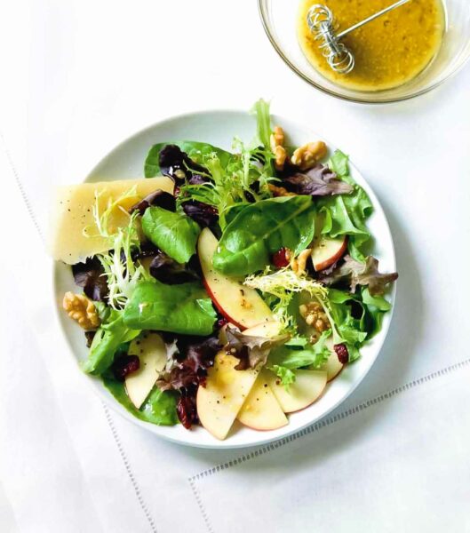 A large white bowl filled with apples, parmesan, walnuts, and mixed green salad, with a small bowl of honey dressing next to it.