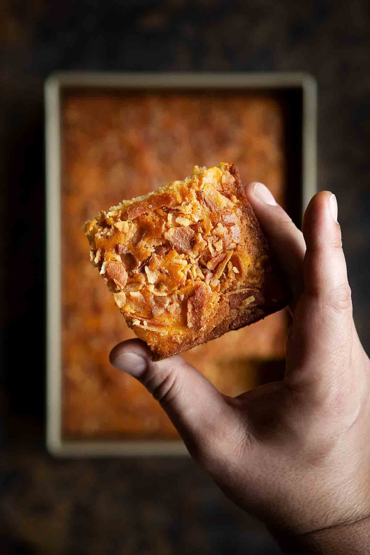 A large rectangular pan with bacon cornbread in the background with a hand holding the corner piece of cornbread.