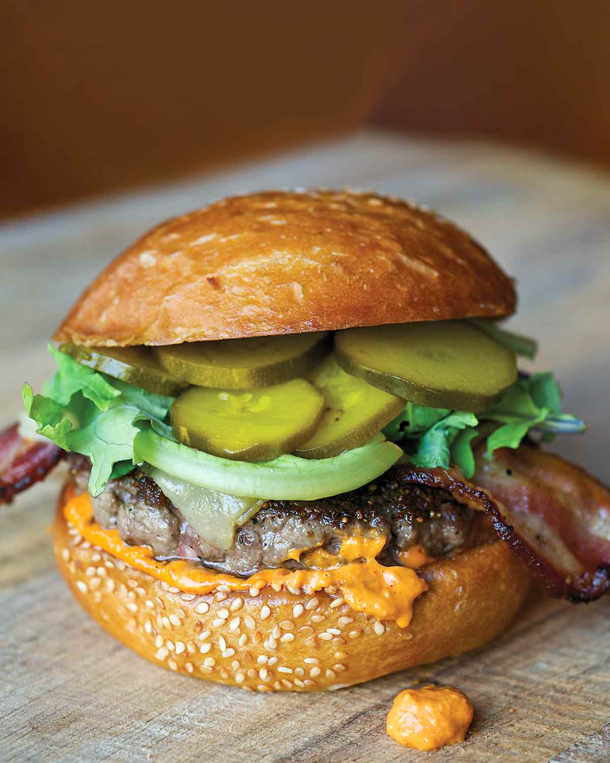 A cheeseburger sitting on a countertop, with a sesame bun, melted cheese, a beef patty, lettuce, bacon, and pickled zucchini.