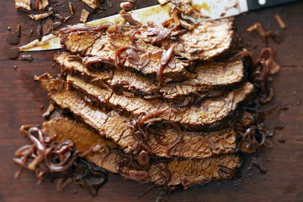 A sliced piece of braised brisket with red wine and honey laying on a cutting board with a large knife and covered with braised onions.