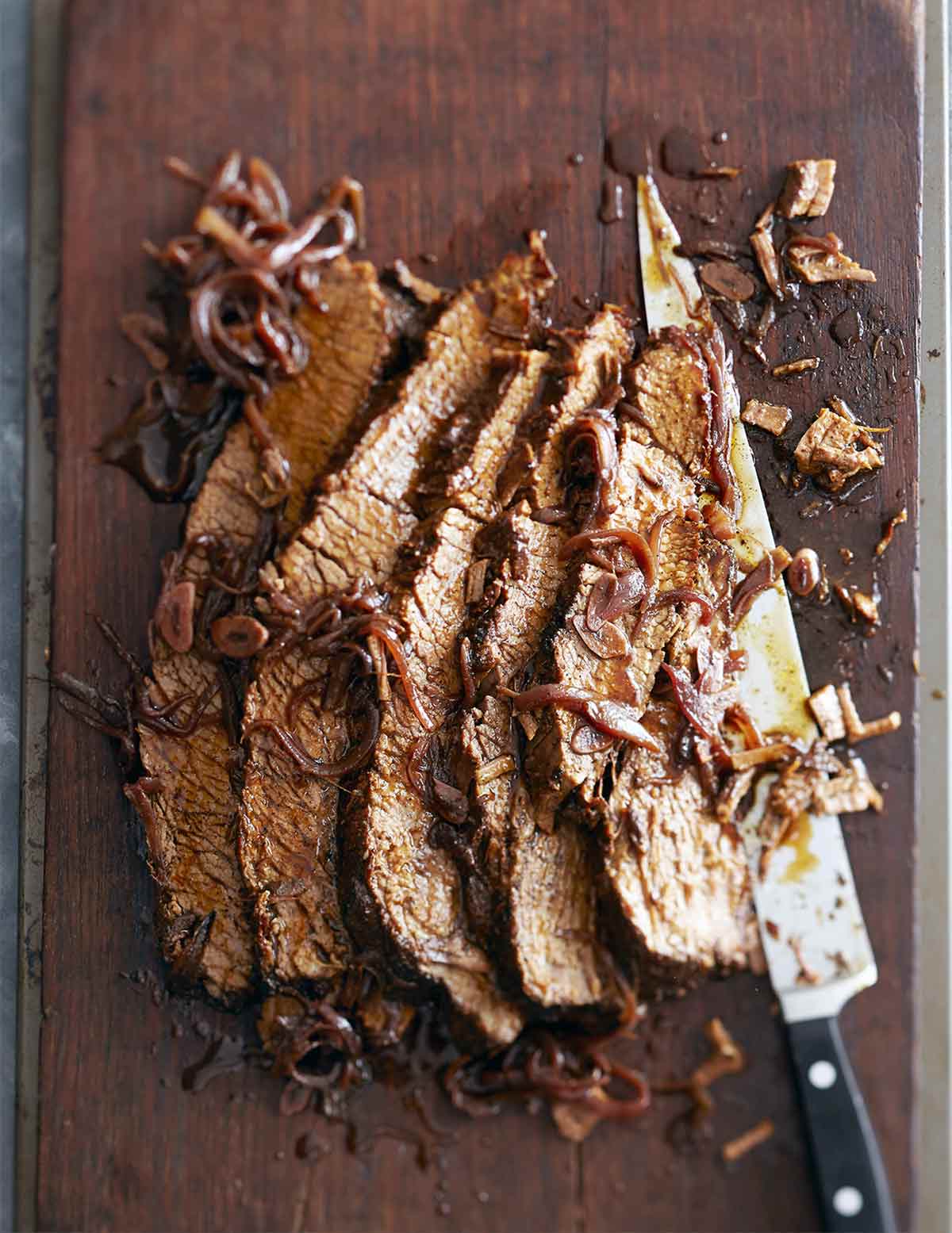 A sliced piece of braised brisket with red wine and honey laying on a cutting board with a large knife and covered with braised onions.