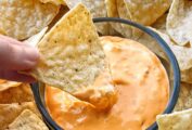 A bowl of butternut queso surrounded by tortilla chips, a hand dipping one into the queso.