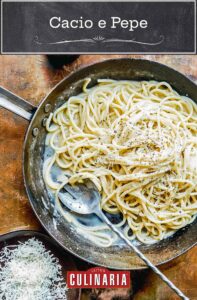 A metal skillet filled with creamy pasta covered with shredded Parmesan and cracked pepper, with a large serving spoon. A bowl of Parmesan and a pepper mill sit beside it.