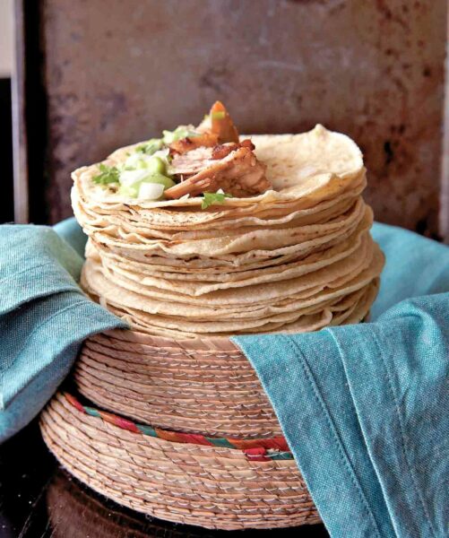 A pile of soft tortillas topped with carnitas, onion, guacamole, and cilantro. They're resting in a woven basket on a pile of blue napkins.