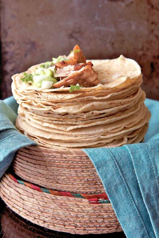 A pile of soft tortillas topped with carnitas, onion, guacamole, and cilantro. They're resting in a woven basket on a pile of blue napkins.