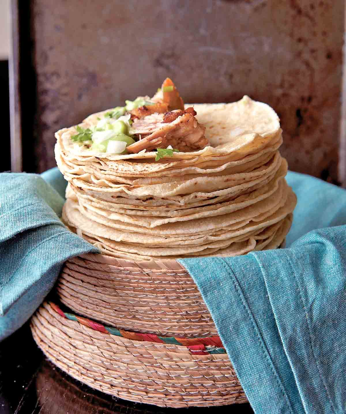En hög med mjuka tortillas toppade med carnitas, lök, guacamole och koriander.  De vilar i en vävd korg på en hög med blå servetter.