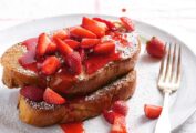 A white plate with 2 pieces of challah French toast covered with fresh strawberries, icing sugar and being drizzled with strawberry syrup.