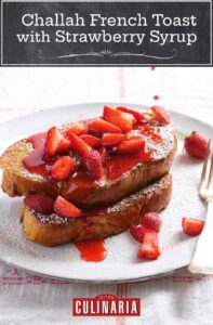 A white plate with 2 pieces of challah French toast covered with fresh strawberries, icing sugar and being drizzled with strawberry syrup.