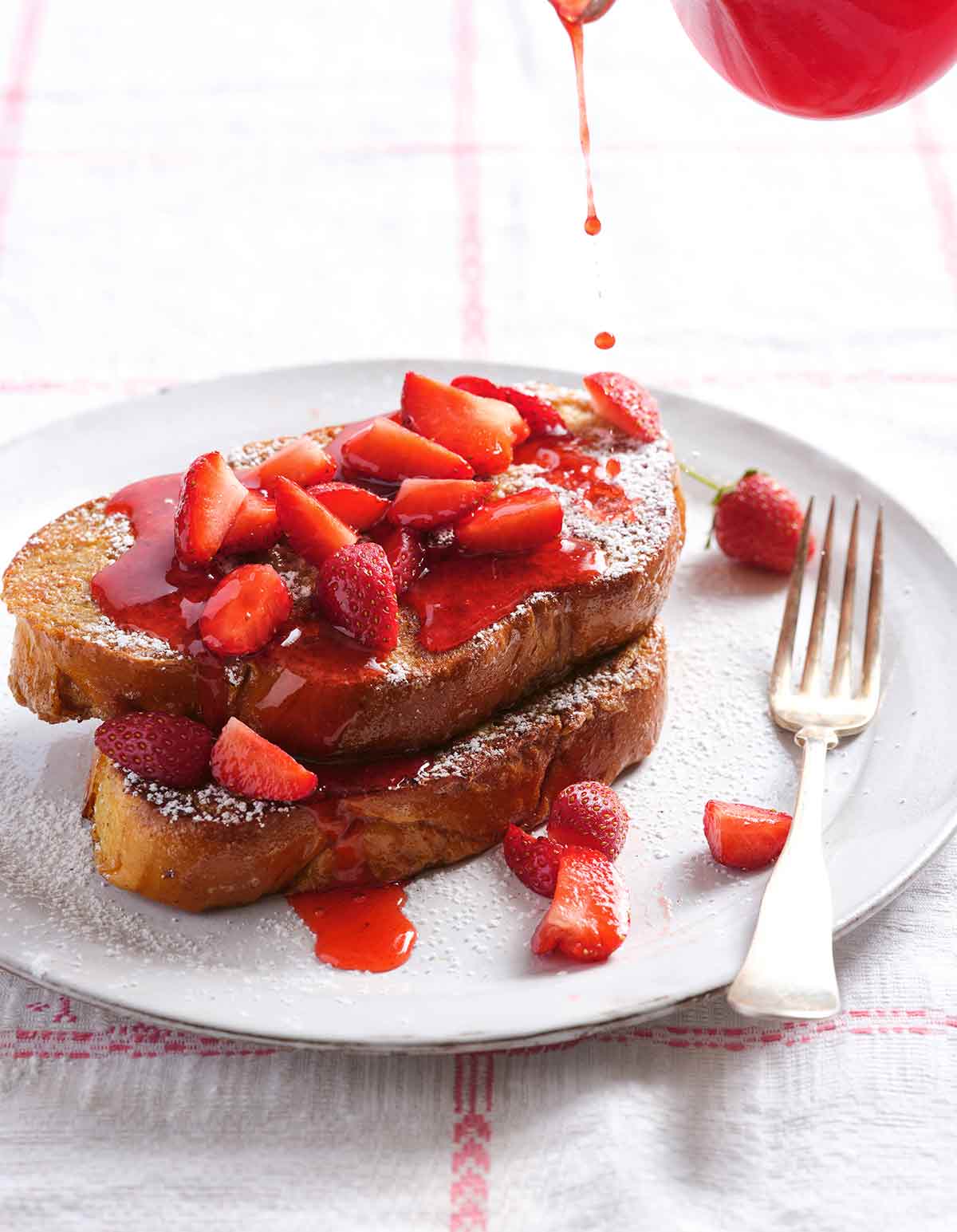 En vit tallrik med 2 bitar challah-franska toast täckt med färska jordgubbar, florsocker och drypps med jordgubbssirap.