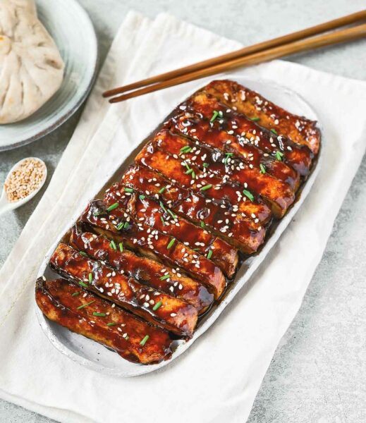A white platter filled with slices of tofu covered in a dark sauce, sesame seeds, and chives, beside a plate of dumplings, a spoon filled with sesame seeds, and a pair of chopsticks.
