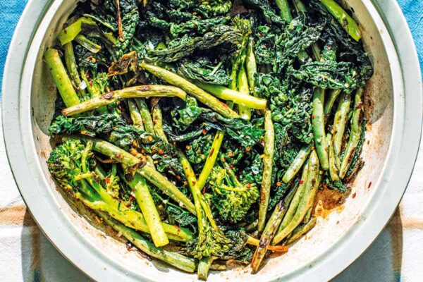 A soft green casserole dish filed with lightly charred broccolini, kale, and green beans, sprinkled with chile flakes and garlic.
