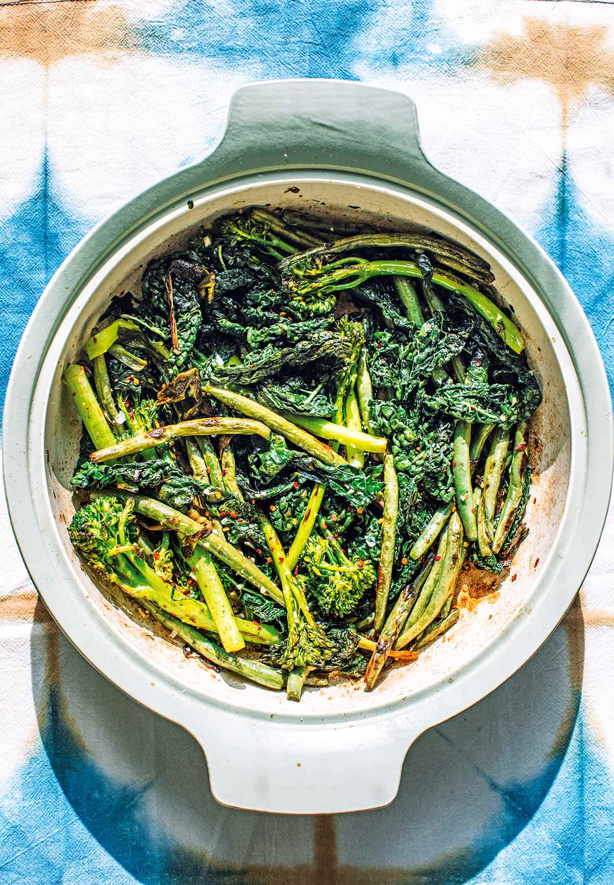 A soft green casserole dish filed with lightly charred broccolini, kale, and green beans, sprinkled with chile flakes and garlic.