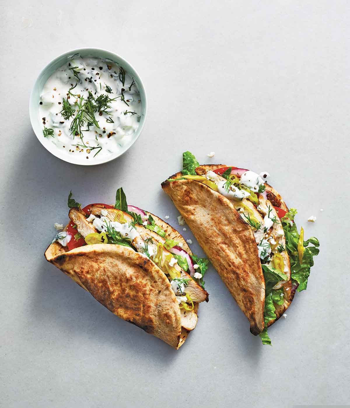 2 chicken gyros filled with romaine, chicken, tomatoes, red onion, pepperoncinis, and tzatziki, with a bowl of tzatziki next to it.