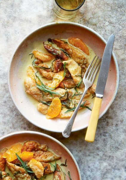 2 bowls of chicken tenders in a cream sauce with slices of orange and tarragon leaves, with a knife and fork.