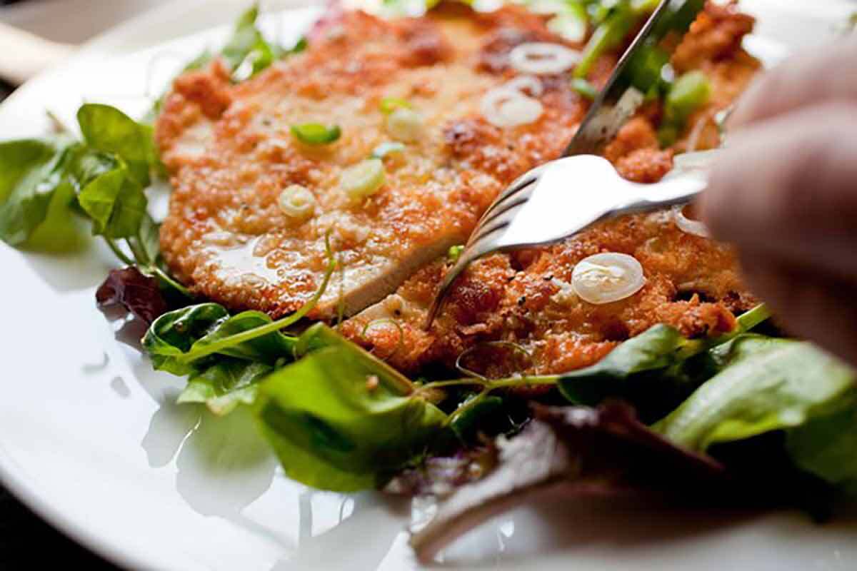 A chicken schnitzel on a white plate with a man's hands cutting into it.