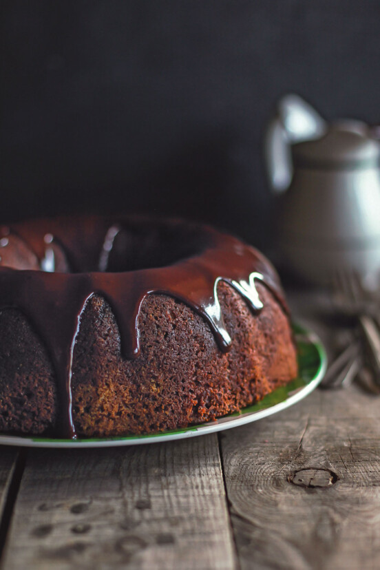 A wooden table with a pitcher and a green plate holding a chocolate bundt cake drizzled in chocolate ganache.