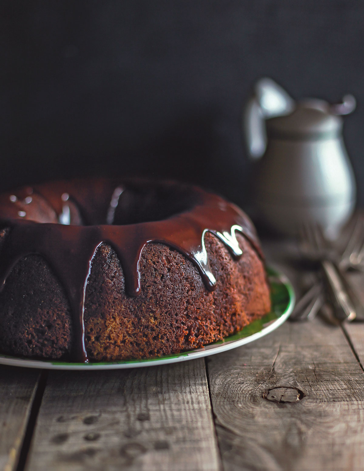 A wooden table with a pitcher and a green plate holding a chocolate bundt cake drizzled in chocolate ganache.