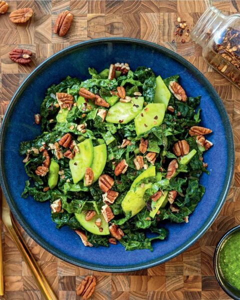 A blue bowl filled with kale, pecans, green apple, and cilantro on a wooden table. A serving spoon, jar of pecans, and bowl of dressing sit next to it.