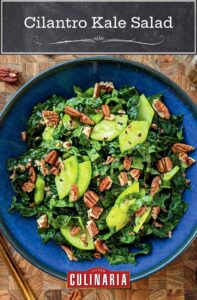 A blue bowl filled with kale, pecans, green apple, and cilantro on a wooden table.