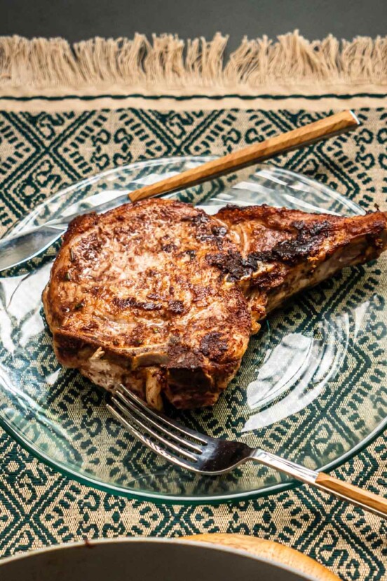A large cinnamon-rosemary pork chop on a glass plate with a knife and fork.
