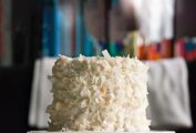 A layered white cake with white frosting and covered with large strips of coconut, sitting on a white cake stand.