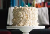 A layered white cake with white frosting and covered with large strips of coconut, sitting on a white cake stand.