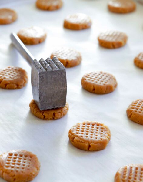 A parchment covered counter with lines of raw peanut butter cookies and a meat-tenderizing mallet.