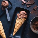 2 rectangular plates with scoops of dark chocolate gelato, one on a scoop and one in a cone, surrounded by squares of dark chocolate and a bowl of cocoa.
