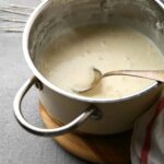 A stainless steel pot, filled with steaming, bubbling easy béchamel sauce with a large spoon and a whisk.
