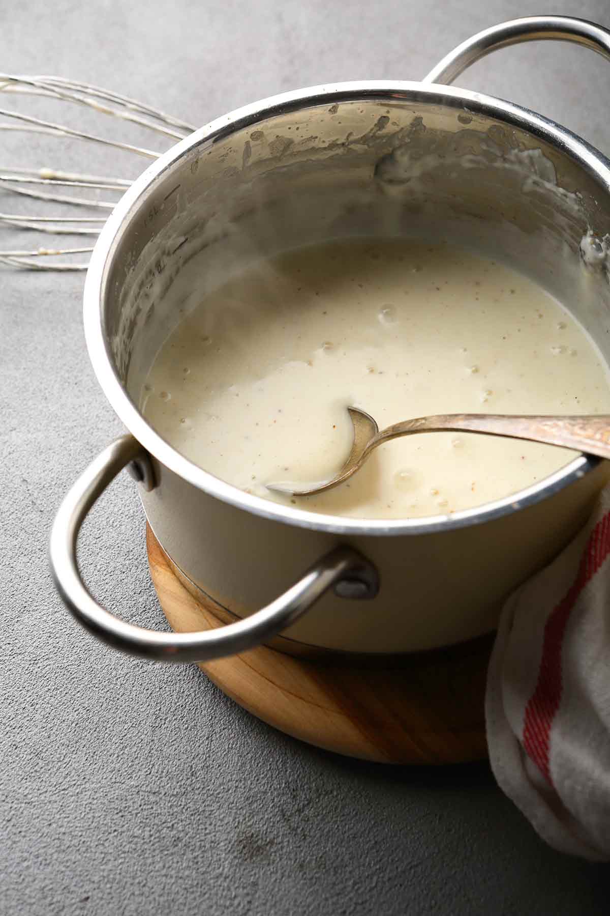 A stainless steel pot, filled with steaming, bubbling easy béchamel sauce with a large spoon and a whisk.