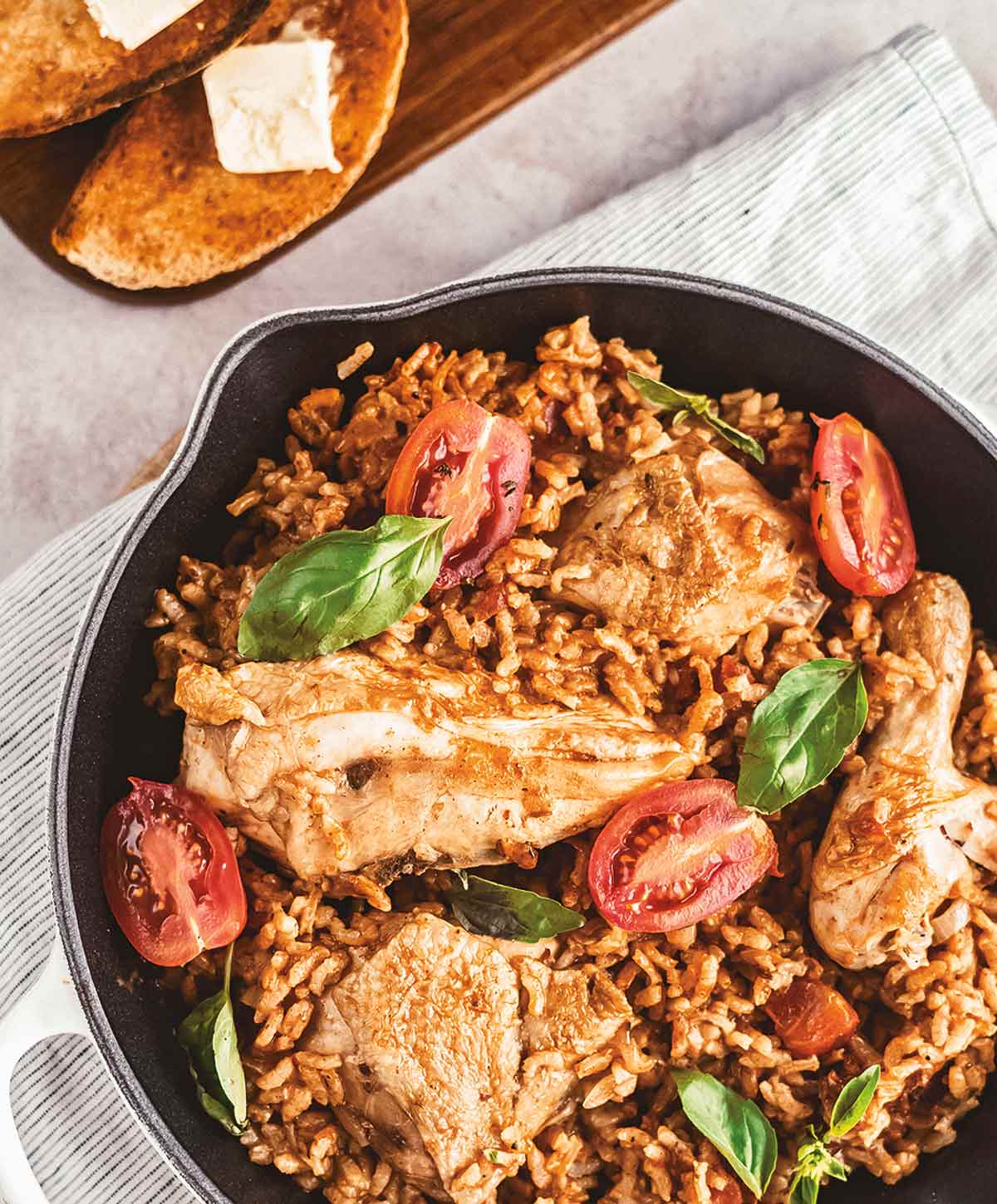A cast-iron skillet with chicken, orzo, tomatoes, and garnished with basil. A wooden bread board with slices of buttered bread sits beside it.
